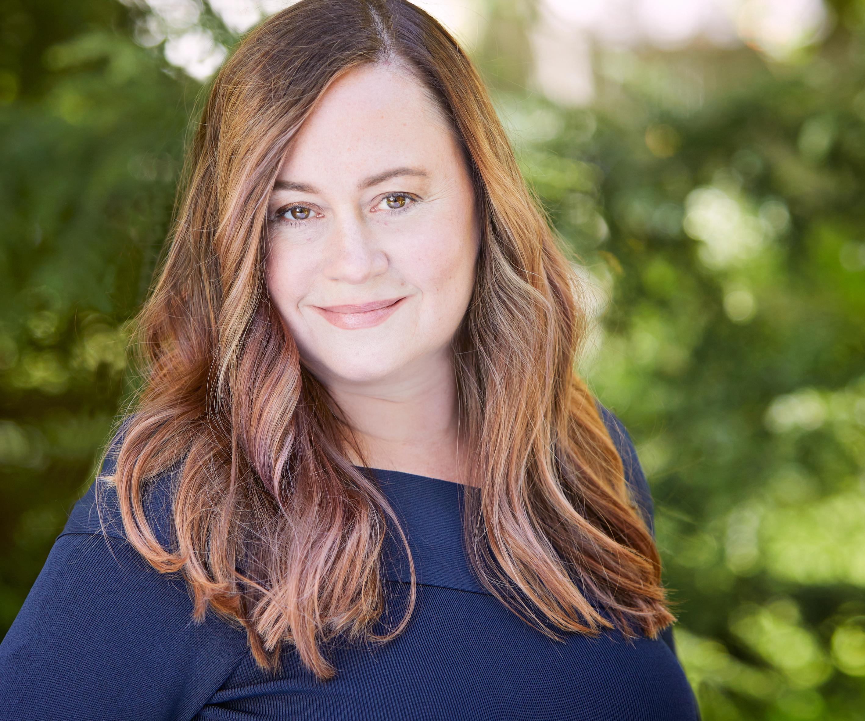 Portrait of author Shelby Van Pelt. A woman smiles softly. Her hair is medium length and wavy, she wears a navy shirt and stands in front of green trees.