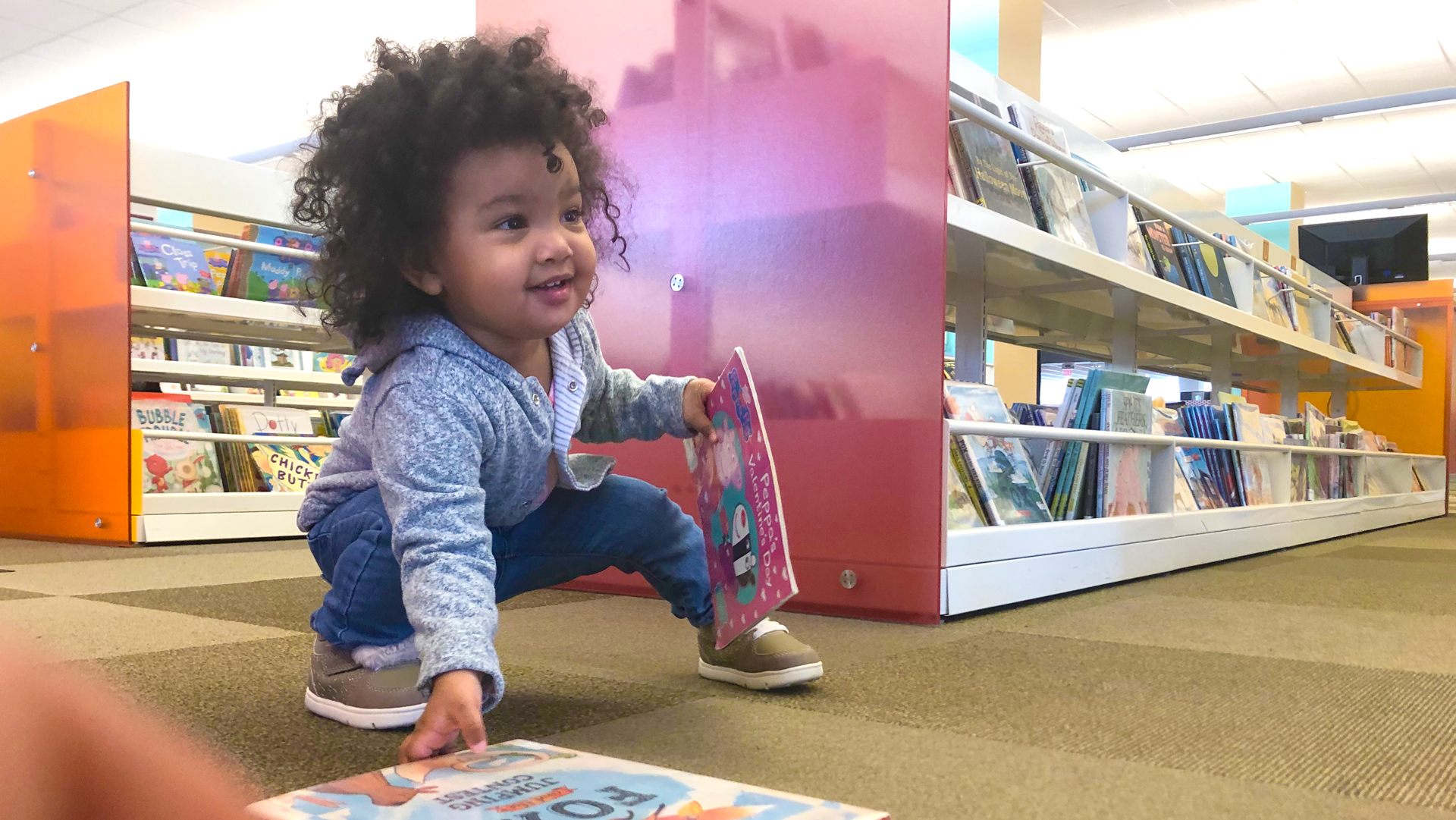 toddler squats, smiles, picks up books, in children's area of library